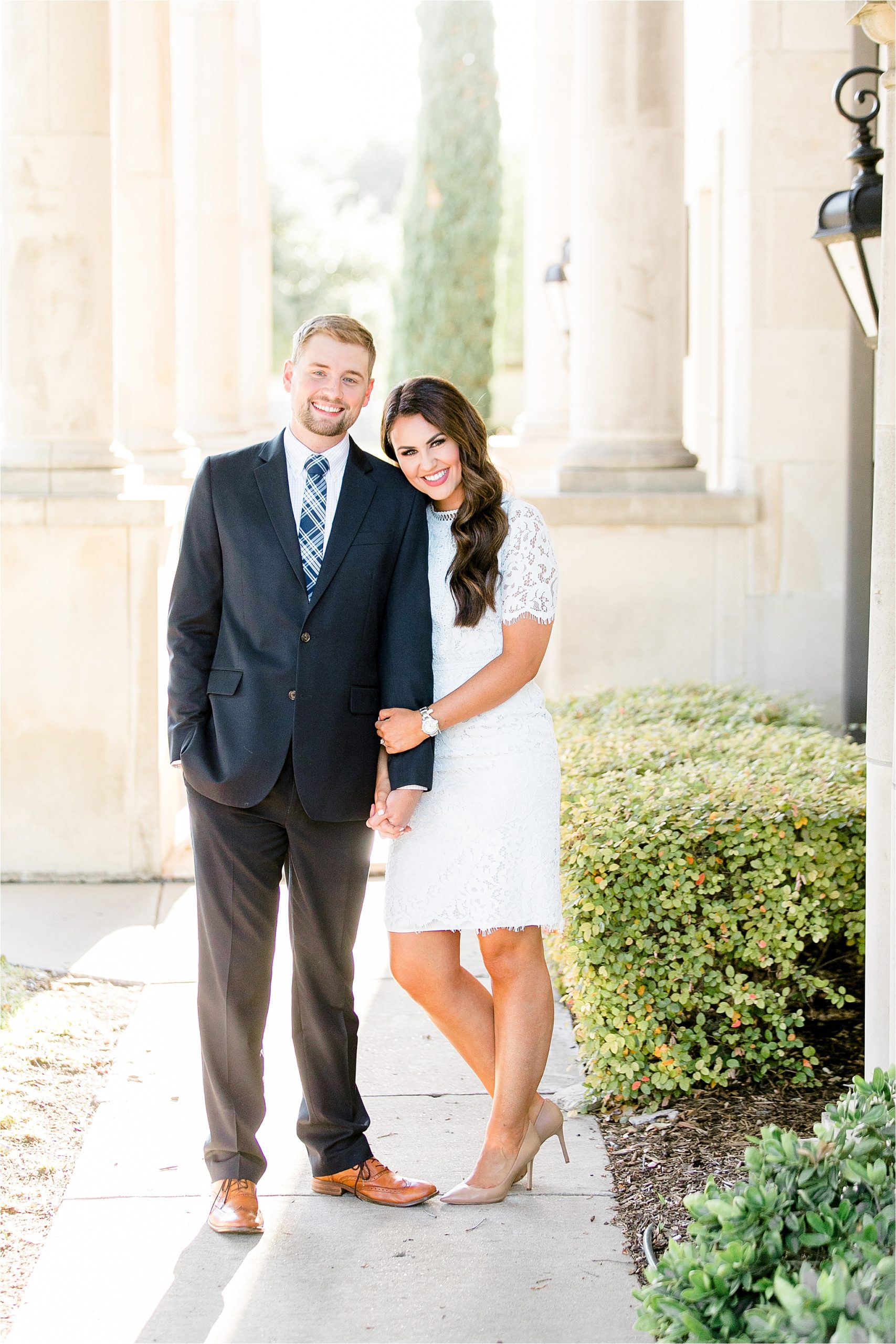 in lace dress leaning on her fiance and holding hands in front of stone columns at Adriatica in McKinney, Texas. 