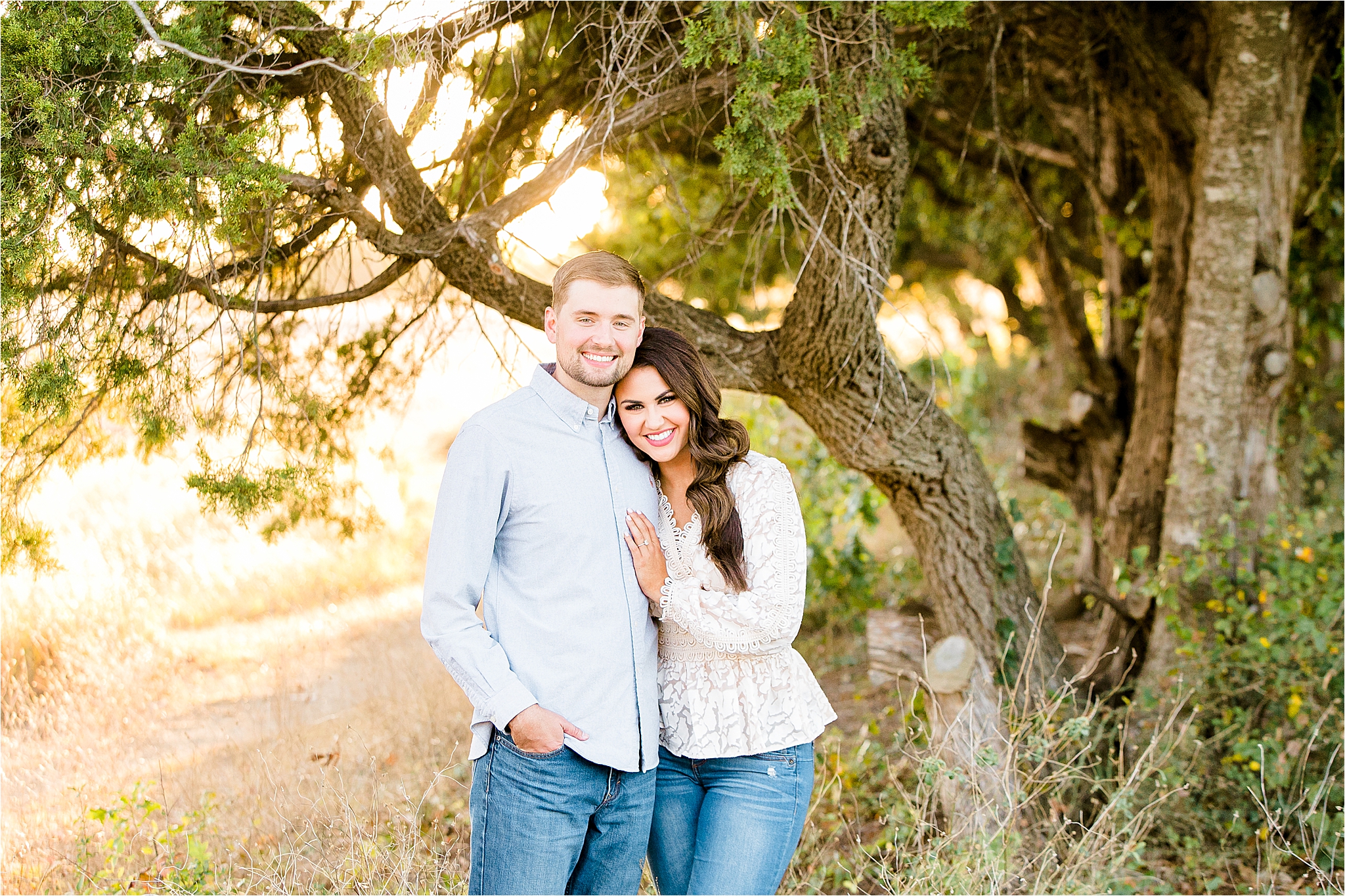 A couple in a field with golden light and trees behind them smiles camera during their engagement session in McKinney, Texas