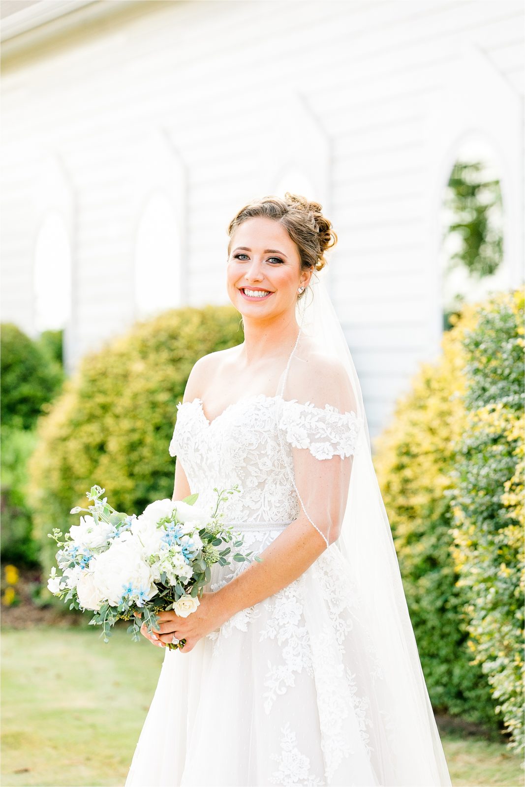 A bride smiles at the camera in an off the shoulder wedding dress and a veil over her shoulders by McKinney Wedding Photographer Jillian Hogan 