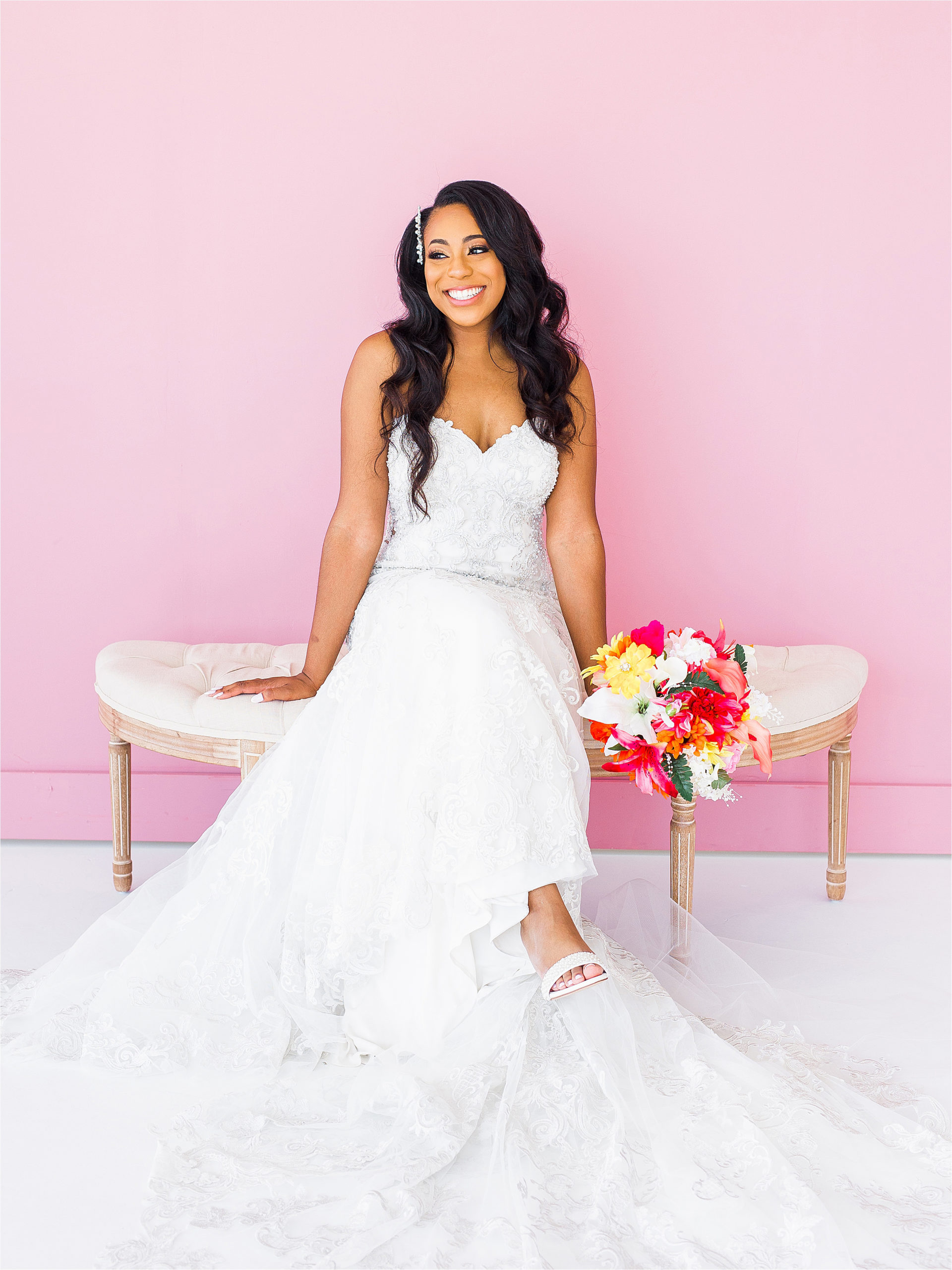 A black bride in a lace dress sits on a white bench in front of a bright pink backdrop with her colorful bridal bouquet next to her and smiling during her san antonio studio bridal portraits 