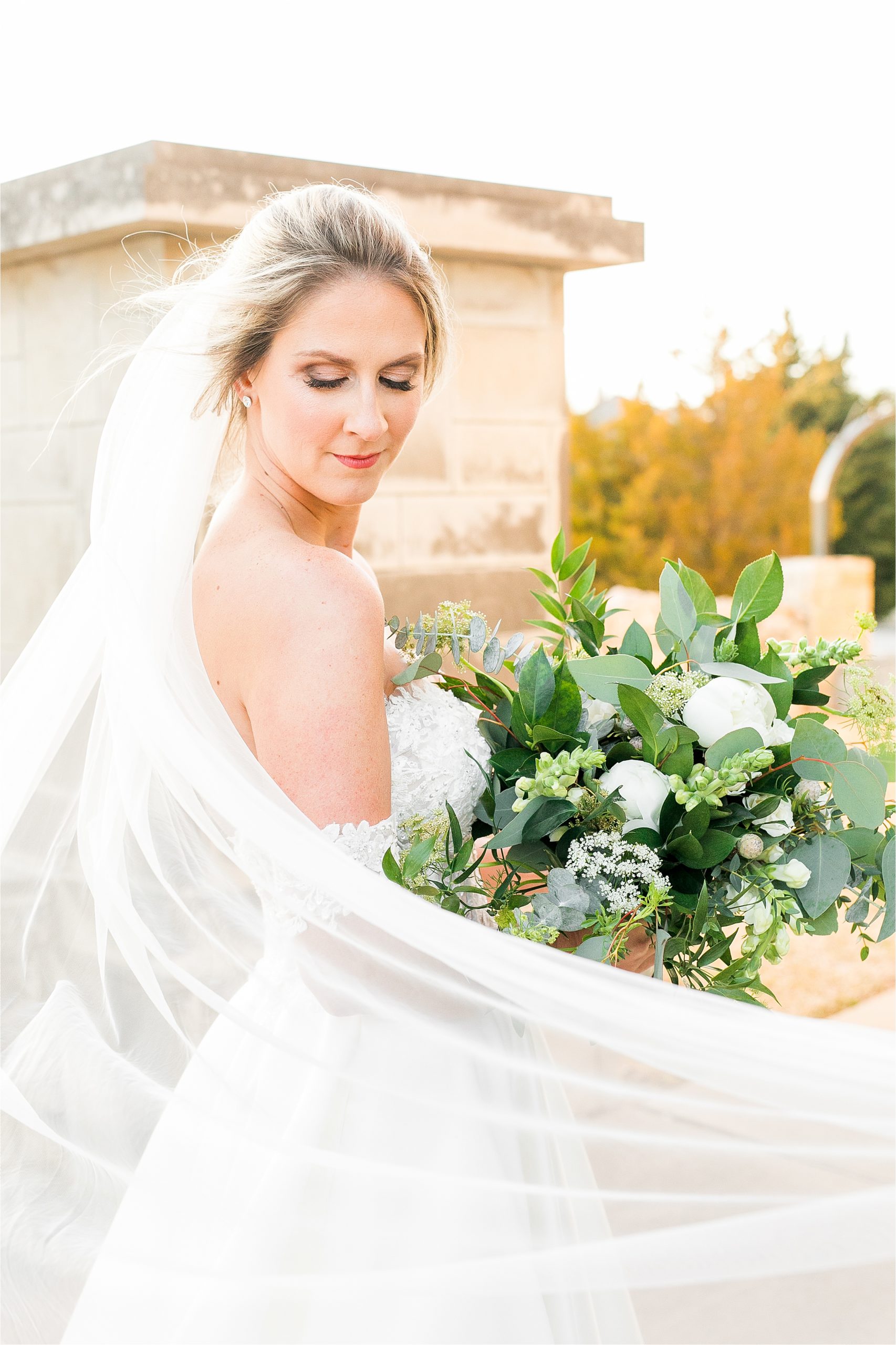 A beautiful bride in a off the shoulder wedding gown leans over her shoulder with her veil swooped in front of her with lots of greenery and lush white flowers at her adriatica village bridal session