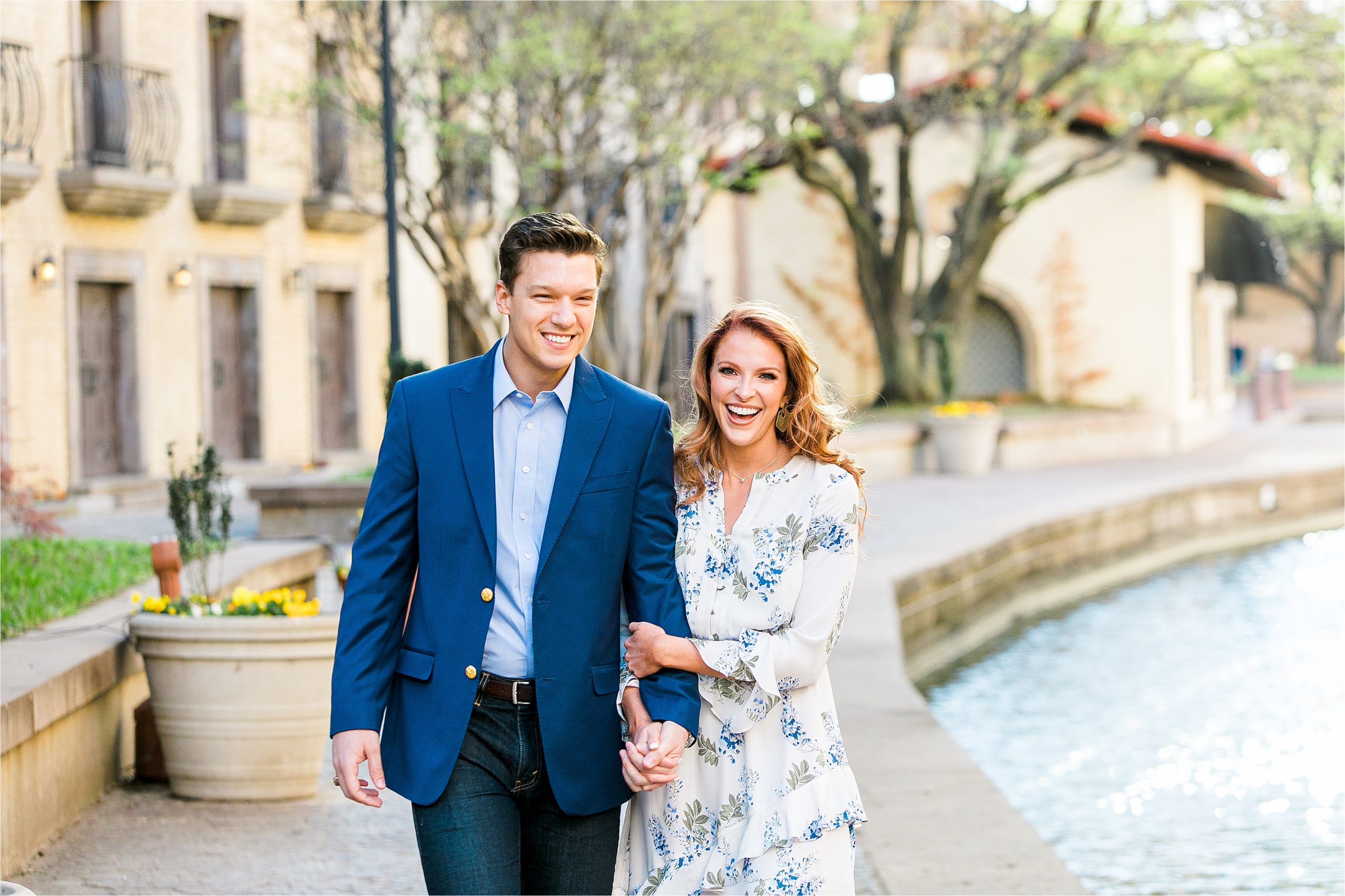 An engaged couple laughs as she grabs his arms and they hold hands strolling along the river with old buildings in the background during their San Antonio Engagement Session with Jillian Hogan Photography 