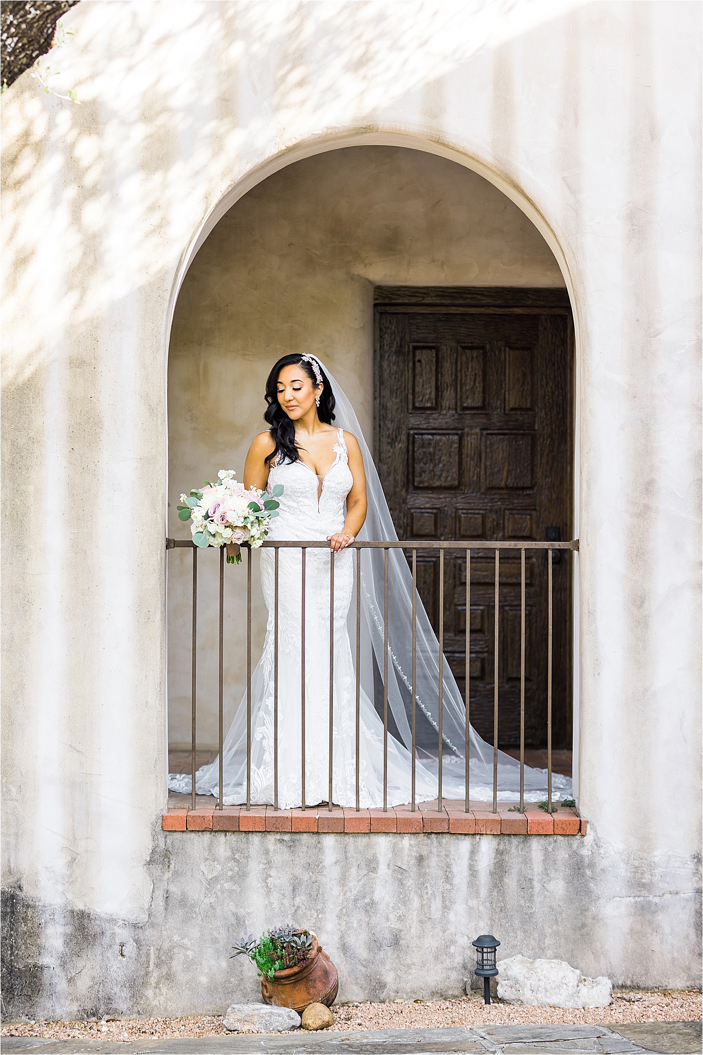A bride in a white lace wedding dress looks down as she holds a railing with a pretty arch over her as her long veil hangs over her shoulders at Lost Mission just outside San Antonio, Texas 