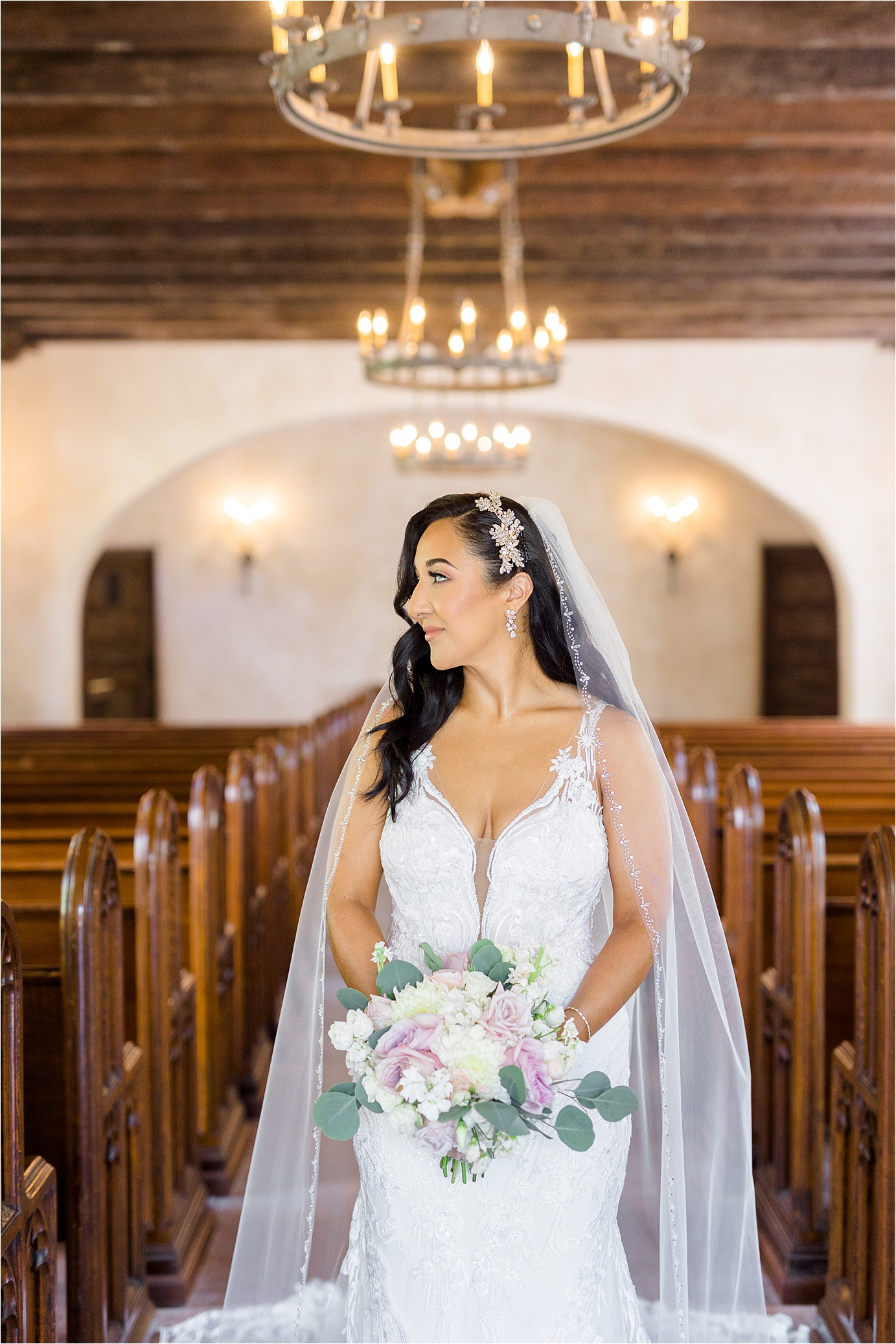 A bride looks over her right shoulder holding a pink and white bouquet with her veil over her shoulders under old chandeliers during her San Antonio Bridal Portraits 