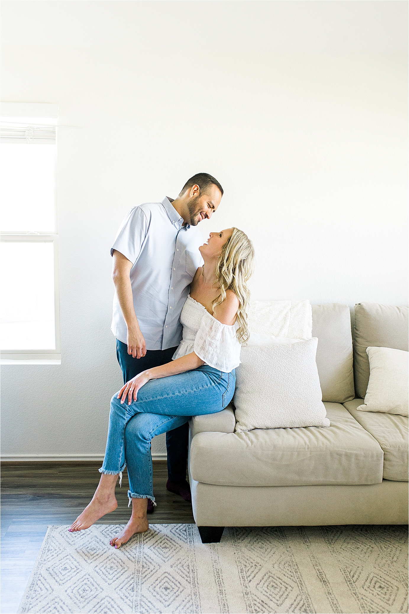 A couple playfully smile at each other while sitting on the edge their couch for an in home engagement session in Austin Texas with photographer Jillian Hogan